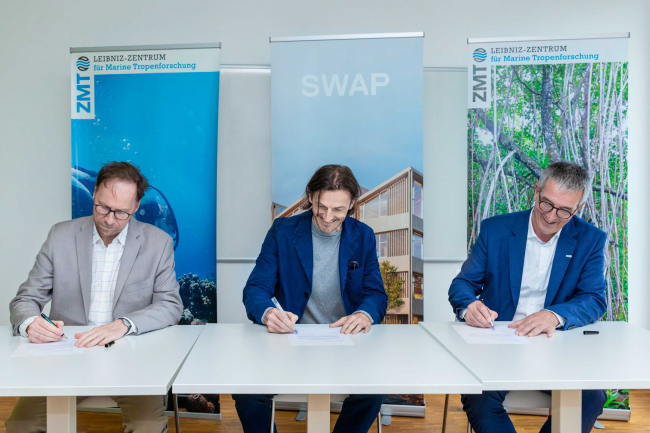Three men sit at a row of tables and sign a paper