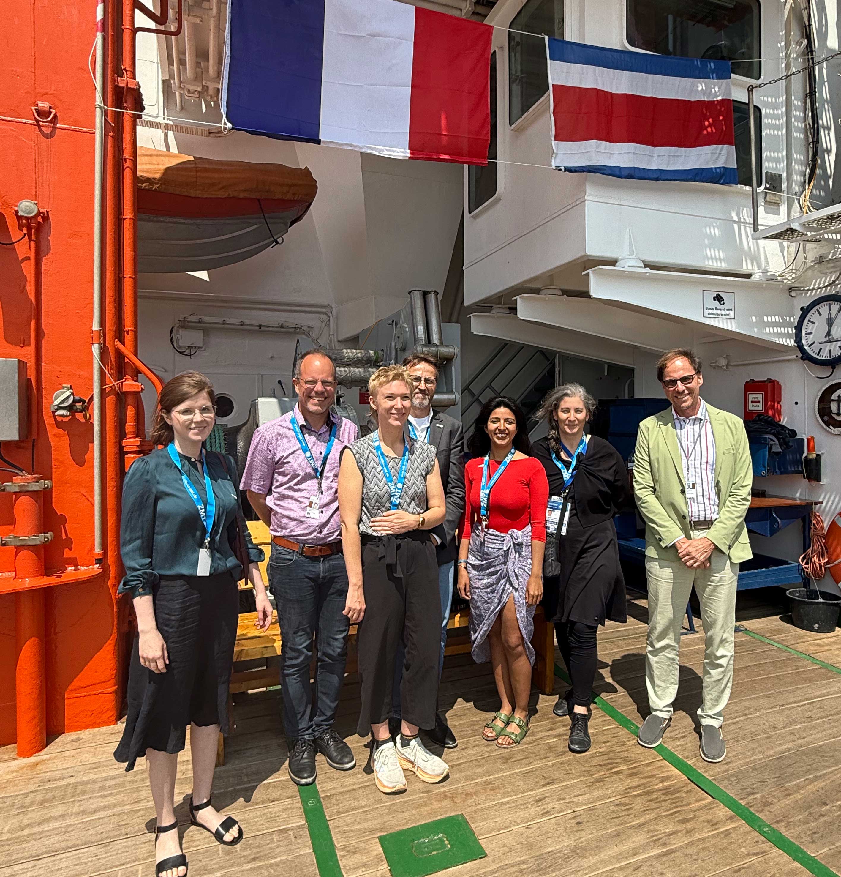 Seven people pose for a group photo on the deck of a research vessel under sunny conditions. They are standing in front of the ship's structure, which features an orange wall and two national flags—France on the left and Costa Rica on the right. The group is dressed in business casual attire with blue lanyards, suggesting participation in an official event. 