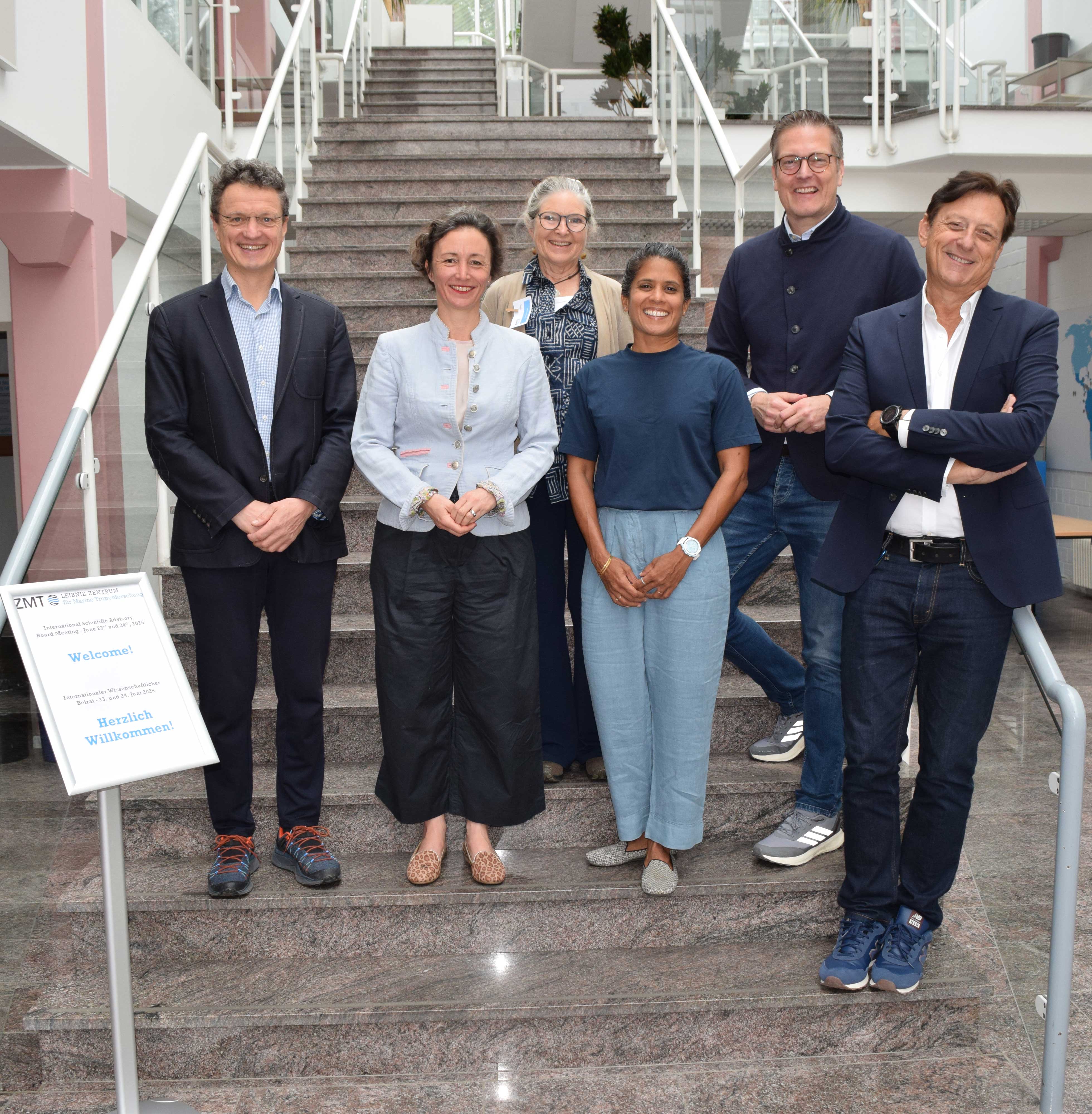 Sechs Erwachsene stehen auf einer Marmortreppe in einem Innenraum und posieren für ein Gruppenfoto. Drei Männer und drei Frauen sind in zwei Reihen angeordnet, wobei die vordere Reihe auf einer niedrigeren Stufe steht. Alle sind leger gekleidet und tragen elegante Freizeitkleidung, darunter Blazer, Hemden mit Knöpfen und lockere Hosen. Sie lächeln und befinden sich in einer professionellen oder akademischen Umgebung zu befinden, mit Glasgeländern und einem modernen Interieur im Hintergrund.