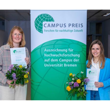 Two women hold certificates in front of a banner.