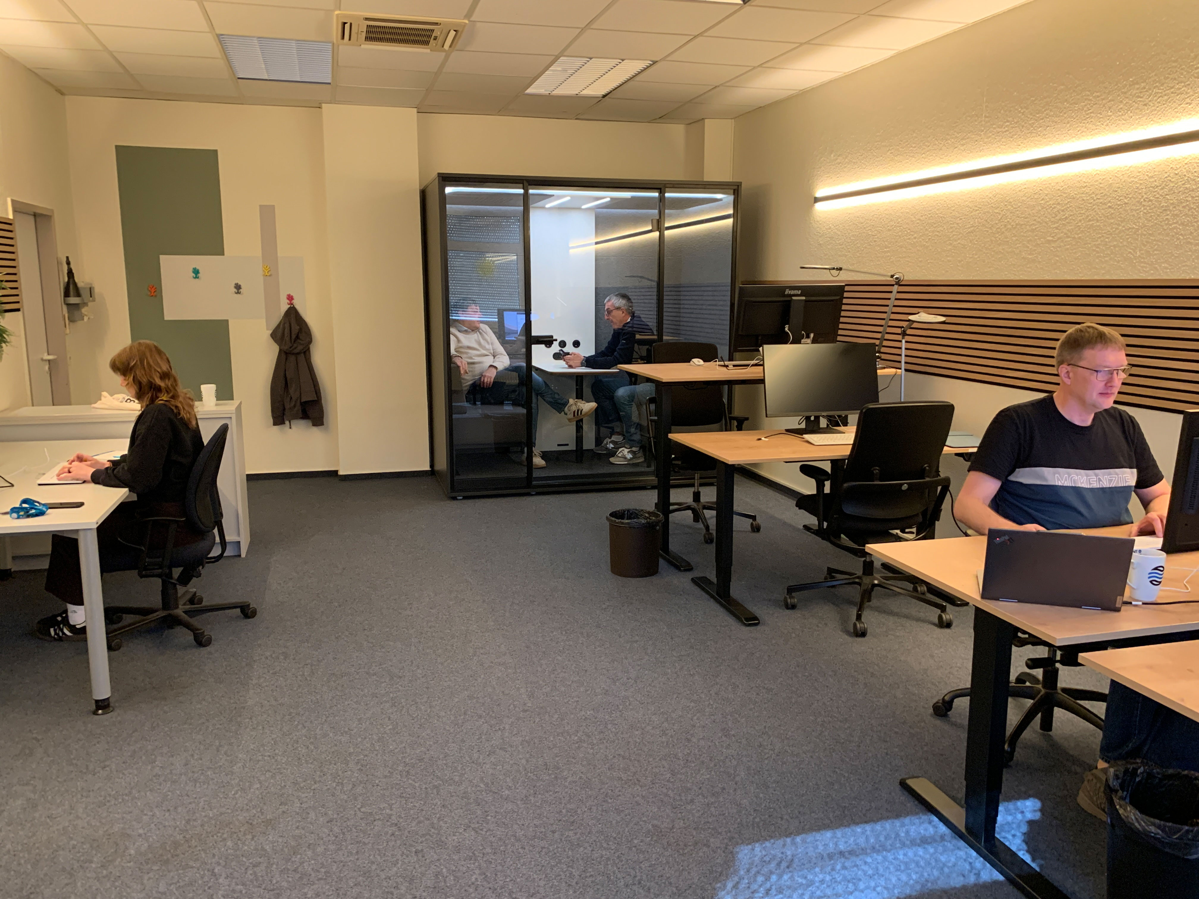 Büro mit hellen Wänden und grauem Teppichboden. Links sitzt eine Frau an einem Schreibtisch und arbeitet am Computer, von der Seite bzw. leicht von hinten zu sehen. Rechts sitzt ein Mann mit Brille an einem separaten Tisch und tippt auf einem Laptop. In der Mitte im Hintergrund befindet sich eine gläserne Meeting-Kabine, in der zwei Personen einander gegenübersitzen und sich unterhalten. Mehrere Schreibtische, Bürostühle, Monitore und ein kleiner Mülleimer sind sichtbar, beleuchtet durch gleichmäßiges Deckenlicht.