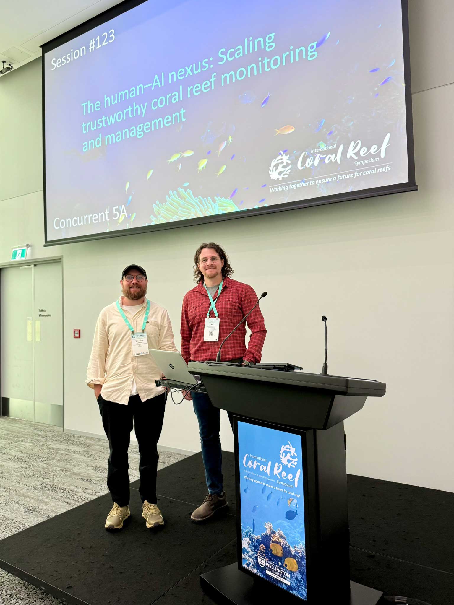 Two men pose for a photo beside a lectern. Above them, a large screen displays the session “The human–AI nexus: Scaling trustworthy coral reef monitoring and management” at the International Coral Reef Symposium.