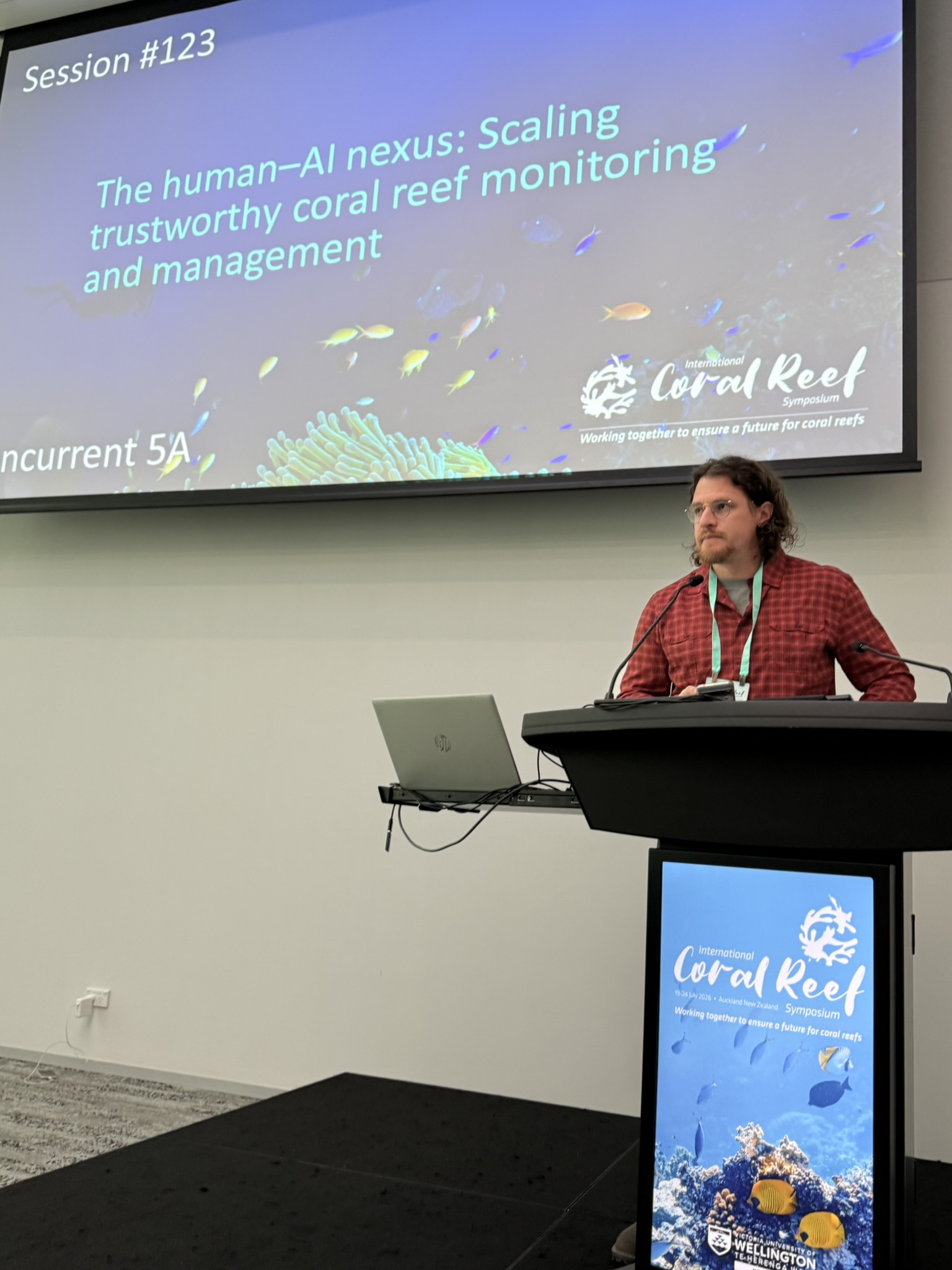 A man speaks at a lectern during a conference session. The large screen behind him displays “The human–AI nexus: Scaling trustworthy coral reef monitoring and management.”