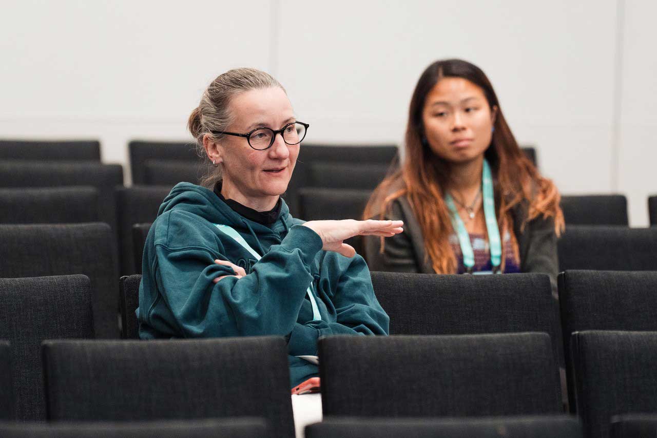 Two people sit in a lecture hall. The person in the foreground gestures with one hand during a conversation, while the second person listens attentively from a few rows behind.