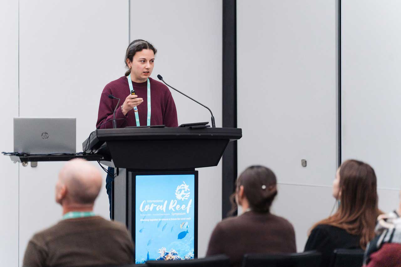 A woman gives a presentation at a lectern, speaking to the audience while holding a presentation remote. Several audience members are visible from behind in the foreground.