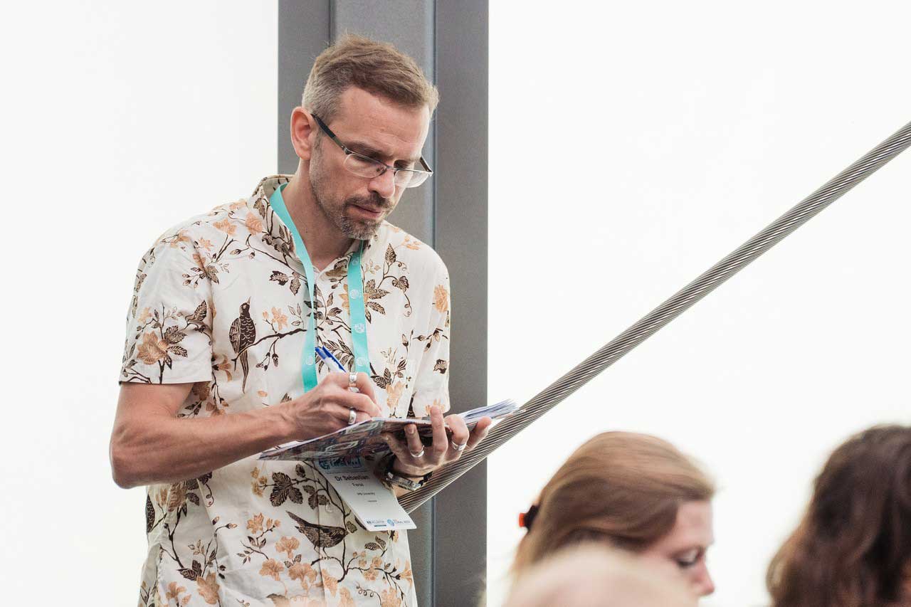 A man stands in an event space, concentrating as he takes notes on a notepad. Other attendees are visible out of focus in the foreground.