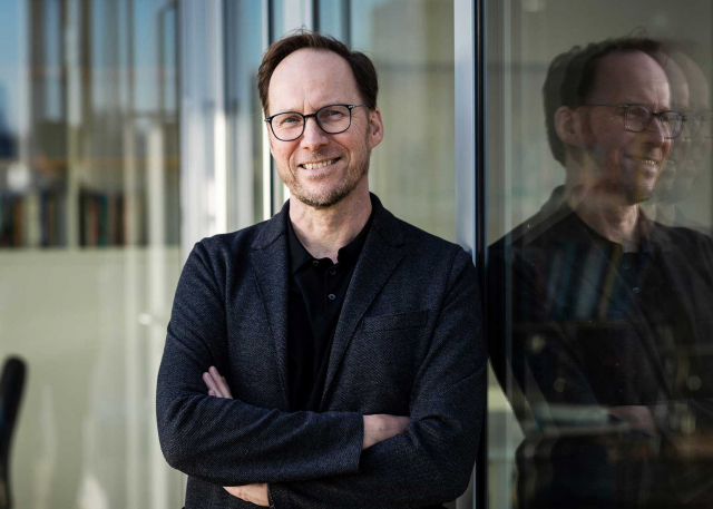 Lächelnder Mann vor einem Fenster, in dem sich das Gesicht des Mannes spiegelt