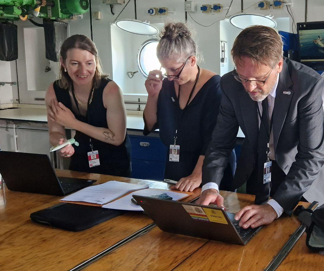 Alt-text: Three people work at a large wooden table inside a ship’s interior room. Two women and one man, all wearing lanyards, are focused on their laptops and documents. The woman on the left is smiling and holding a pen, the middle woman is adjusting her glasses while reading, and the man on the right is typing on a laptop. The room has round porthole windows, overhead machinery, and industrial equipment, indicating a working space aboard a research vessel.