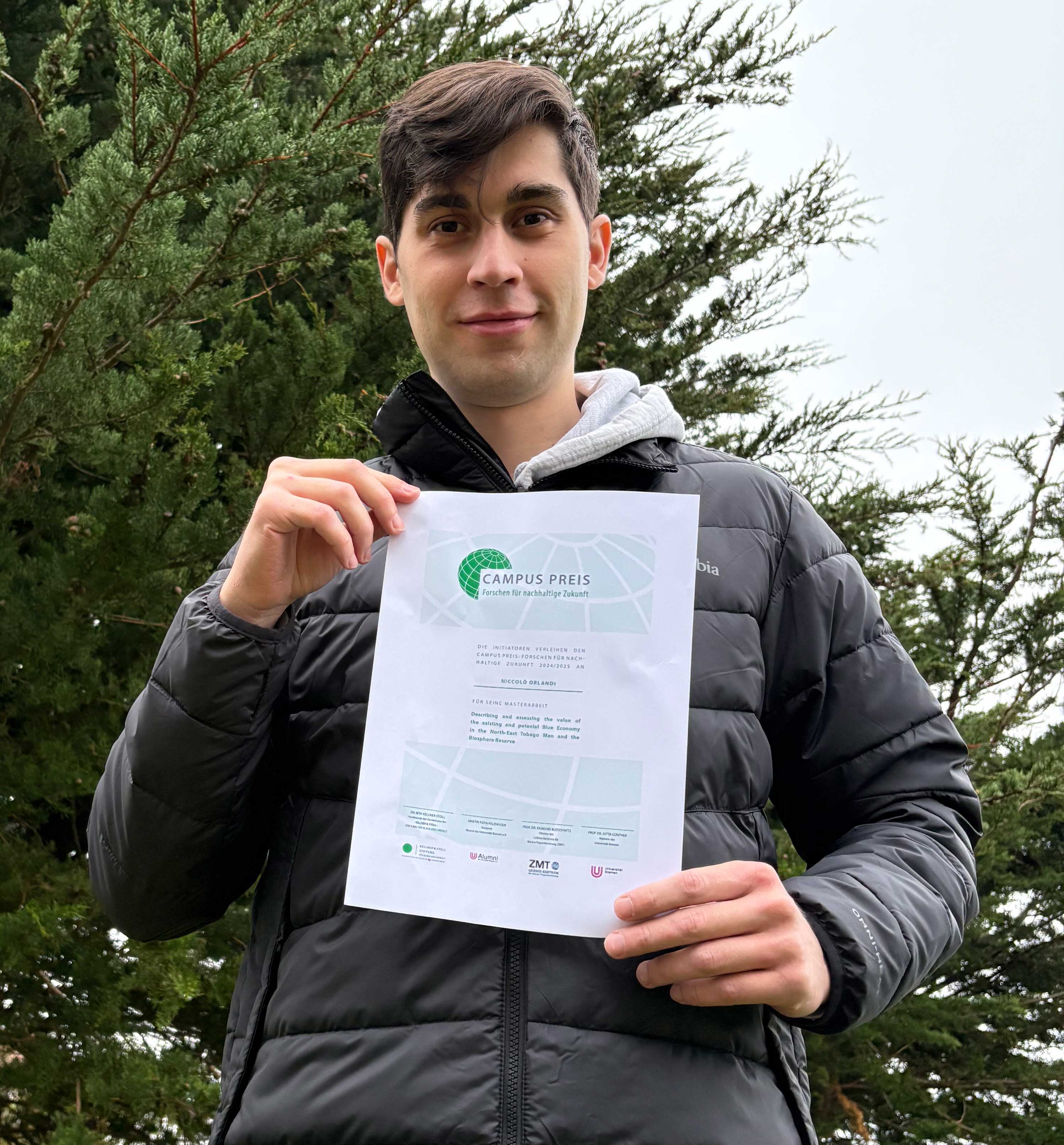 A young man in a black jacket holds up a “Campus Preis” certificate while standing outdoors in front of green trees.