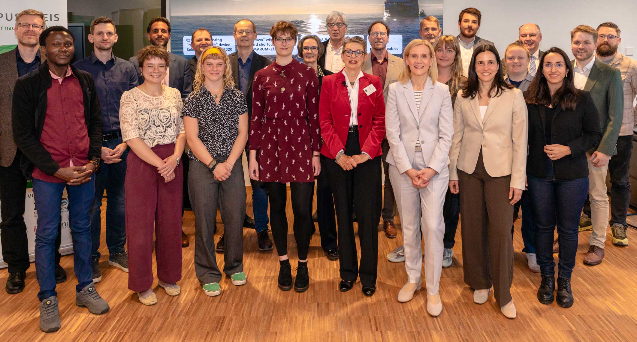Large group photo of about twenty researchers and professionals standing indoors on a wooden floor in front of a large presentation screen. The group includes men and with several people smiling at the camera. 