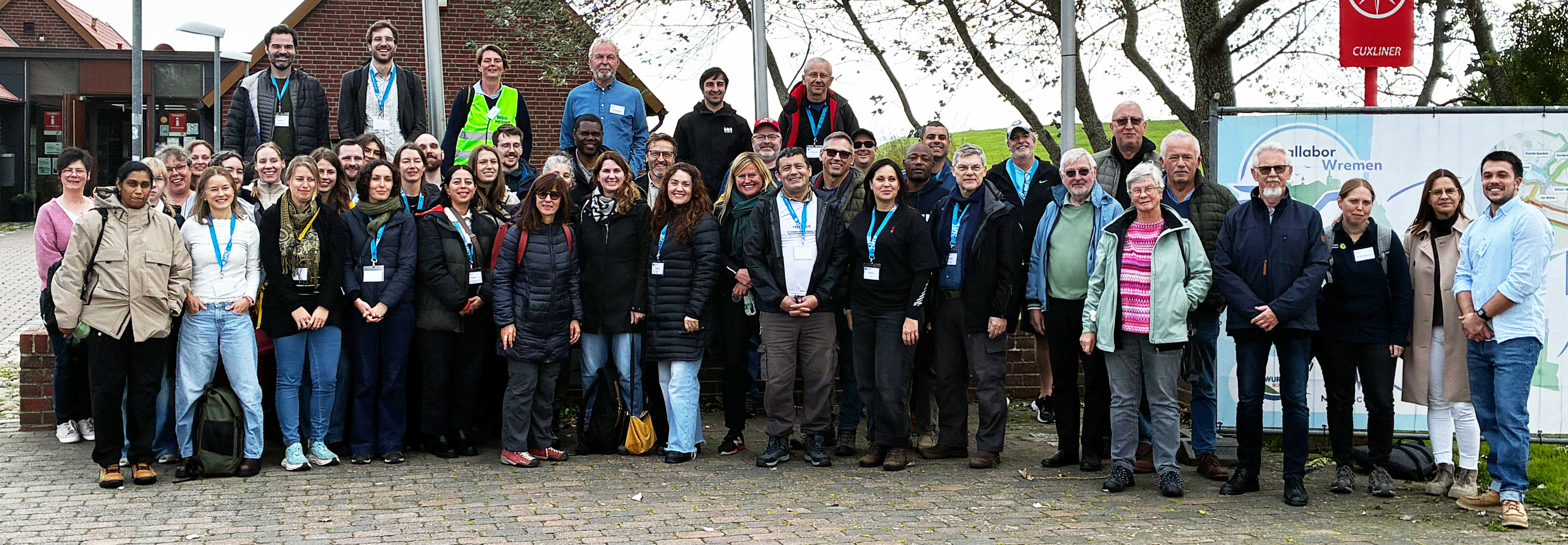 Große Gruppe von Teilnehmenden posiert gemeinsam für ein Gruppenfoto im Freien. Im Hintergrund stehen ein Backsteingebäude, Fahnenmasten und Bäume.