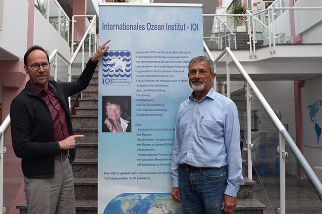 two men in front of a roll-up banner, the man on teh right points at writing on the banner