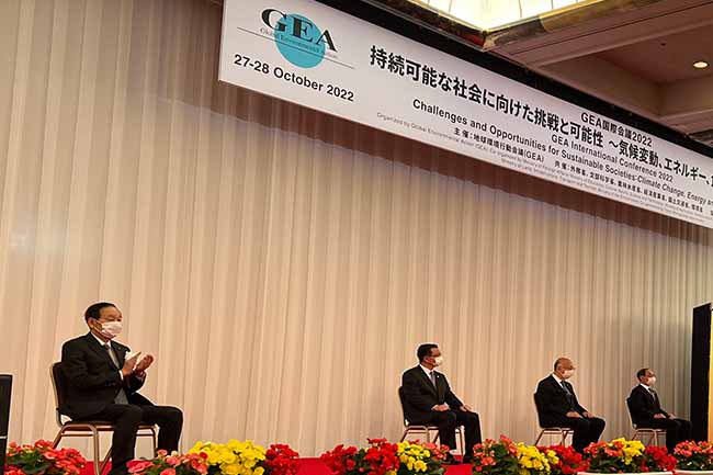 Four men in black suits sit on a stage which is decorated with flowers in the front