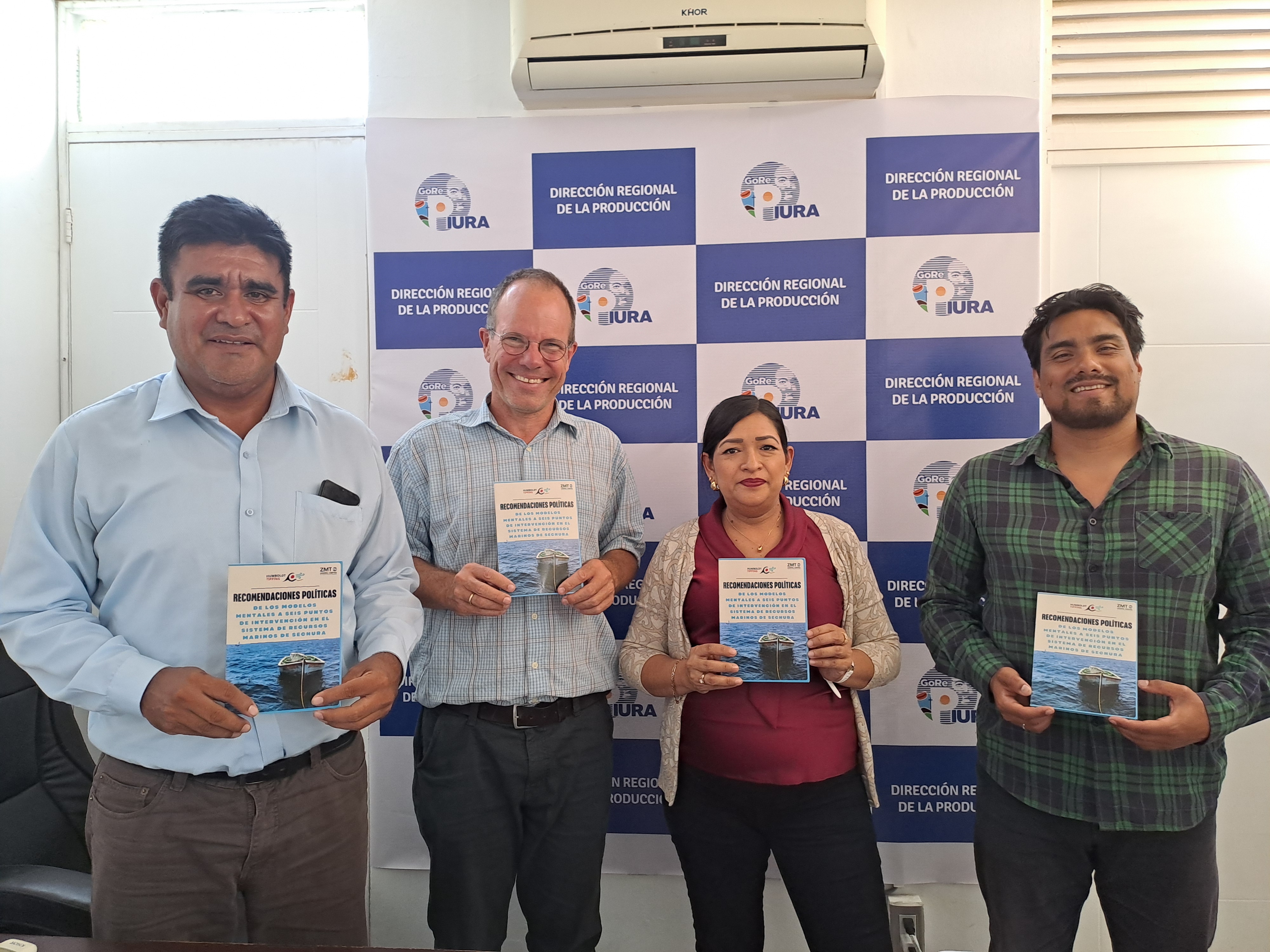 Four people stand side by side in an office, each holding the same publication titled "Recomendaciones Políticas". The cover features a small boat on the ocean. They are posing in front of a press backdrop displaying the logo of "Dirección Regional de la Producción" and the text "GORE PIURA". All four individuals are smiling and looking at the camera.