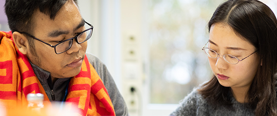 wo people sit side by side at a table, focused on reading or writing. The person on the left wears glasses, a gray jacket, and a bright red-and-orange patterned scarf draped over their shoulders. The person on the right also wears glasses and a gray fuzzy sweater. Both appear to be concentrating, with their heads slightly tilted downward.