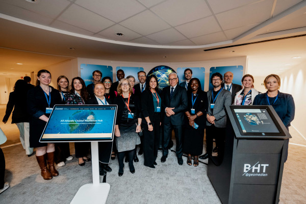 A diverse group of around 20 people stands together indoors, smiling for a group photo in front of a digital display and a globe graphic. A diverse group of around 20 people stands together indoors, smiling for a group photo in front of a digital display and a globe graphic.