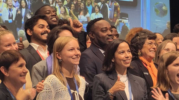A large, diverse group of people stands closely together, smiling and celebrating at an indoor event. Many individuals are giving thumbs-up gestures and appear joyful and enthusiastic. A large, diverse group of people stands closely together, smiling and celebrating at an indoor event. Many individuals are giving thumbs-up gestures and appear joyful and enthusiastic.