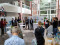 A group of people are gathered in a sunlit indoor atrium for a presentation or discussion. The audience stands in a semicircle around a few tall cocktail tables, attentively listening to a woman speaking near a set of scientific posters. A group of people are gathered in a sunlit indoor atrium for a presentation or discussion. The audience stands in a semicircle around a few tall cocktail tables, attentively listening to a woman speaking near a set of scientific posters.