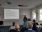 A woman is giving a presentation in a well-lit conference room with large windows. She stands at a podium with a laptop, gesturing as she speaks to an audience seated in front of her. A woman is giving a presentation in a well-lit conference room with large windows. She stands at a podium with a laptop, gesturing as she speaks to an audience seated in front of her.