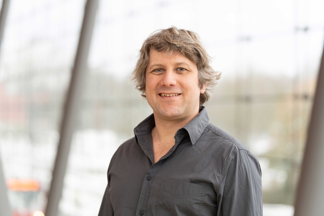 Ein lächelnder Mann mit welligem, hellbraunem Haar und blauen Augen steht in einem Innenraum nahe großer Fenster. Er trägt ein dunkelgraues Hemd mit offenem obersten Knopf. Der Hintergrund ist unscharf und zeigt eine helle, moderne Umgebung mit hohen Glasfronten und Metallstreben.