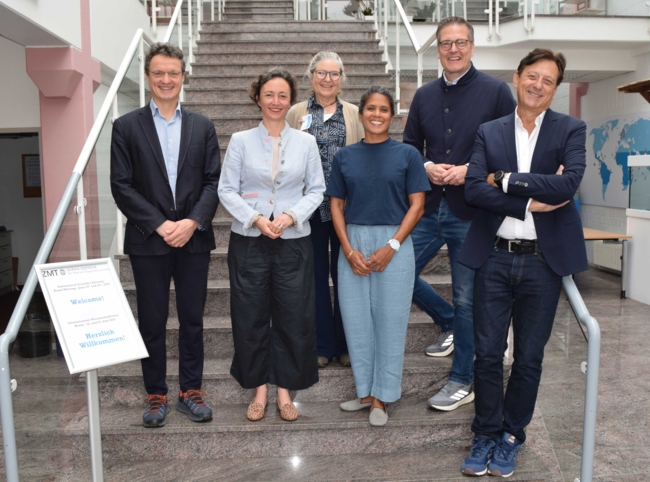 Sechs Erwachsene stehen auf einer Marmortreppe in einem Innenraum und posieren für ein Gruppenfoto. Drei Männer und drei Frauen sind in zwei Reihen angeordnet, wobei die vordere Reihe auf einer niedrigeren Stufe steht. Alle sind leger gekleidet und tragen elegante Freizeitkleidung, darunter Blazer, Hemden mit Knöpfen und lockere Hosen. Sie lächeln und befinden sich in einer professionellen oder akademischen Umgebung zu befinden, mit Glasgeländern und einem modernen Interieur im Hintergrund.