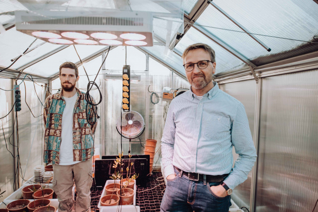 Two men standing in glasshouse, one on the left with dark hair and beard, the other on the right  wearing glasses, jeans and pale blueshirt, photo by Ben Eichler
