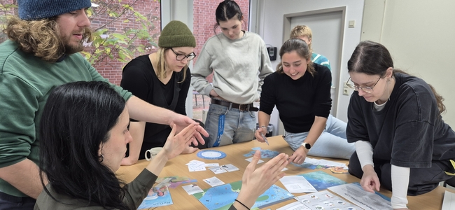 A group of seven adults gathers around a table covered with blue cards, printed sheets, and circular diagrams. One person gestures with both hands while others lean in, looking down at the materials. The group appears engaged in discussion or a collaborative activity in a bright room.