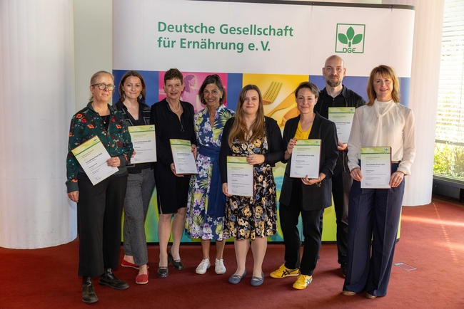 Group photo of eight people standing indoors on a red carpet, holding certificates in front of a backdrop with the logo and name of the "Deutsche Gesellschaft für Ernährung e. V." (German Nutrition Society).