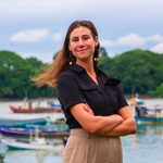 Eine Person mit verschränkten Armen steht lächelnd am Wasser. Im Hintergrund sind mehrere Boote sowie eine grüne Uferlandschaft unter bewölktem Himmel zu sehen.