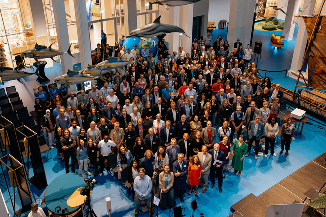 Gruppenfoto aus der Vogelperspektive von etwa 200 Personen, die sich in einem großen Museum oder Wissenschaftszentrum mit hellblauem Boden versammelt haben. Die Menschenmenge, bestehend aus Erwachsenen unterschiedlichen Alters und unterschiedlicher Herkunft, blickt in die Kamera und füllt den größten Teil der zentralen Bodenfläche aus. Über den Köpfen hängen mehrere große Modelle von Meerestieren, darunter Delfine und thunfischähnliche Fische, die den Eindruck erwecken, über den Besuchern zu schwimmen. Rechts ist ein großes Holzboot oder Kanu zu sehen, während im Hintergrund glaswandige Ausstellungsräume und Sitzbereiche zu erkennen sind.