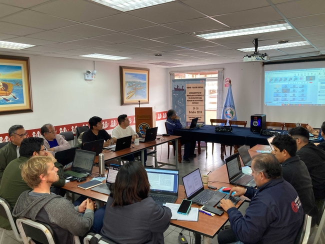 People sit around a U-shaped table in a conference room, each focused on a laptop.  A projector screen on the right displays a software interface, and the room is decorated with paintings of boats.