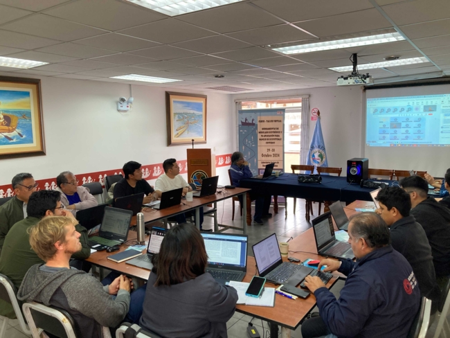 People sit around a U-shaped table in a conference room, each focused on a laptop.  A projector screen on the right displays a software interface, and the room is decorated with paintings of boats.