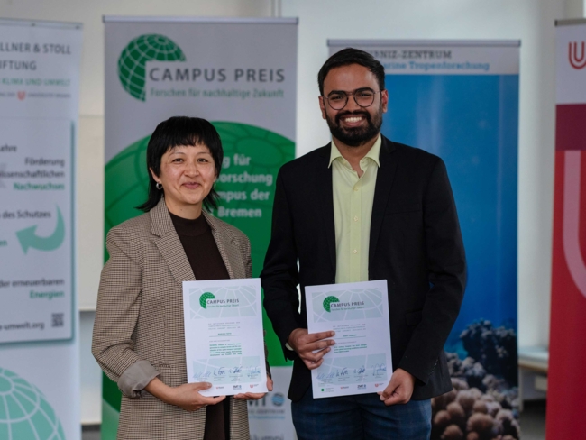 Two people stand side by side indoors, smiling and holding certificates in front of several promotional banners. The woman on the left has short black hair and wears a brown plaid blazer over a dark top. The man on the right has a beard, glasses, and is dressed in a black blazer over a light green shirt. Both certificates are labeled “CAMPUS PREIS – Forschen für nachhaltige Zukunft,” indicating an award related to sustainable research. The background includes logos and banners for environmental and research organizations.