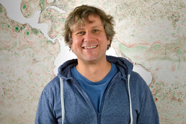 Ein lächelnder Mann mit welligem, hellbraunem Haar steht vor einer farbenfrohen, abstrakten Karte. Er trägt einen blauen Zip-Hoodie über einem blauen T-Shirt. Die Karte im Hintergrund zeigt wirbelnde grüne, rote und beige Muster, die an topografische oder ozeanografische Daten erinnern.