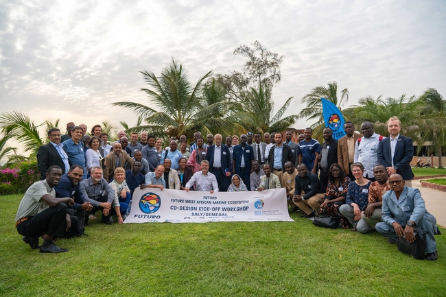 Eine große Gruppe von Menschen posiert für ein Foto im Freien auf einer Rasenfläche, umgeben von Palmen und unter einem bewölkten Himmel. Die Gruppe besteht aus vielfältigen Männern und Frauen unterschiedlichen Alters, die überwiegend in business-casual oder formeller Kleidung gekleidet sind. Vorne wird ein weißes Banner gehalten, auf dem steht: „FUTURO – Future West African Marine Ecosystem – Co-Design Kick-Off Workshop – Saly/Senegal.“ Am unteren Rand des Banners befinden sich die Logos verschiedener unterstützender Organisationen. Rechts in der Gruppe wird eine hellblaue Fahne mit dem FUTURO-Logo gehalten.