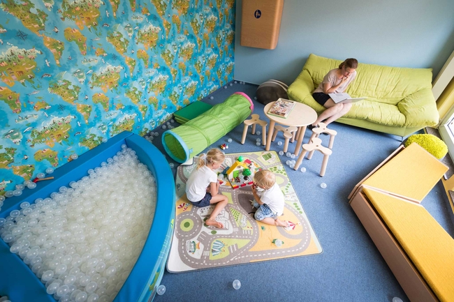Kinder spielen in einem Spielzimmer mit Spielzeug und einem Sofa. Im Hintergrund arbeitet eine Frau an einem Laptop.