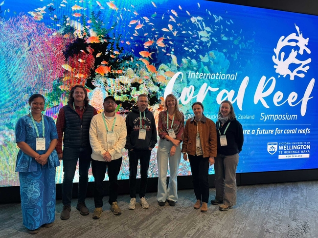 Seven people stand side by side for a group photo in front of a large, colorful screen showing a coral reef with numerous fish. The screen displays the words “International Coral Reef Symposium