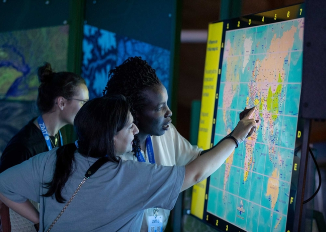 Mia-Sophie Specht (MPI-M), Parima Hajializadeh (ZMT), and Abbie Allela (NAM fellow, Kenia) Three women stand closely together examining a large, brightly lit world map displayed vertically on a screen. Two of the women are actively pointing at a region on the map, appearing engaged in discussion or learning. The map is gridded with labeled coordinates along the top and sides.