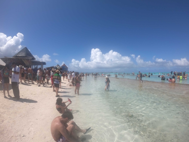 Eine belebte tropische Strandszene mit Dutzenden von Menschen, die einen sonnigen Tag genießen. Viele stehen oder waten im klaren, seichten türkisfarbenen Wasser, während andere am Sandstrand entlanggehen oder sich ausruhen