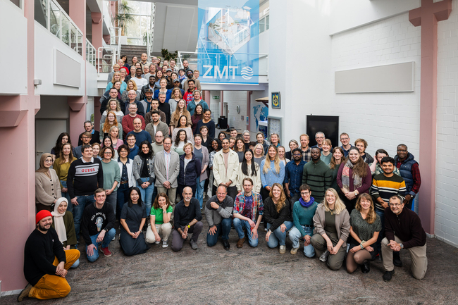 Das Foto zeigt eine große Gruppe von Menschen, die sich im Fyoer eine Gebäudes für eien Foto aufgestellt haben. Sie lächeln in die Kamera.