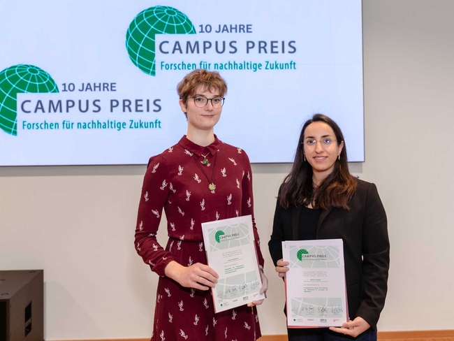 Two women stand side by side holding award certificates in front of a large screen displaying the “10 Jahre Campus Preis – Forschen für nachhaltige Zukunft” logo. The person on the left wears glasses and a long-sleeved burgundy dress with a white bird pattern, while the person on the right wears glasses, a black blazer, and a black top. Both face the camera and smile slightly in an indoor event setting.
