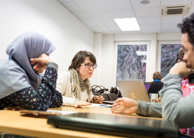 Four people sit around a table in a brightly lit room, engaged in discussion. A woman in glasses and a beige scarf is speaking with an expressive hand gesture, while the others listen attentively. Laptops, notebooks, and pens are on the table.