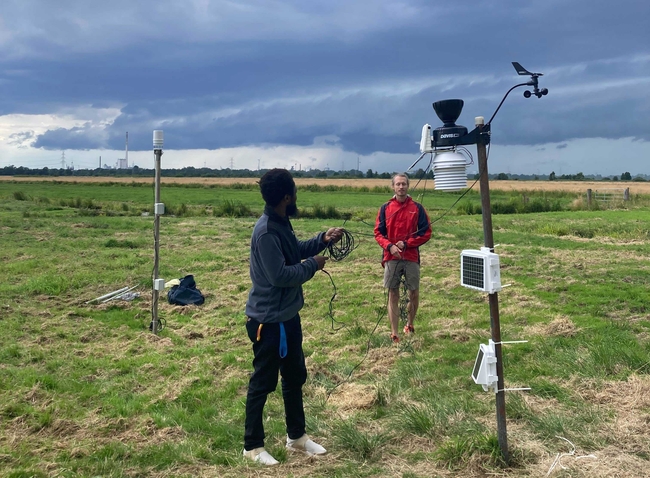 Zwei Personen stellen Wetterstation auf in einem grasbewachsenen Feld unter dunklen Gewitterwolken, wobei am fernen Horizont Industriegebäude zu sehen sind.